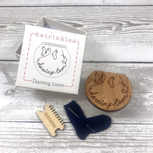 Katrinkles Darning Loom kit box and its contents: a darning loom, heddle, and tapestry needle tucked into a sock shaped piece of felt.