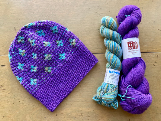 Purple hat with variegated blue and green flowers lying next to two skeins of yarn on a wooden surface.
