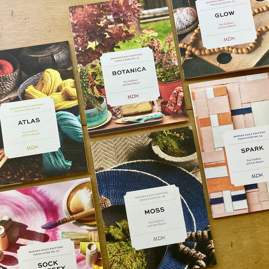 Six different Modern Daily Knitting Field Guides arranged on a wooden surface.