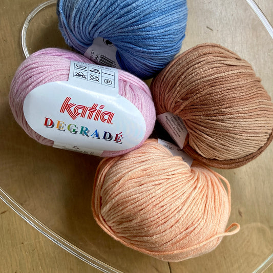 Four colorful balls of Katia Degrade yarn in a clear bowl on a wooden surface.