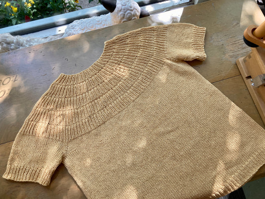 Pale yellow handknit tshirt lying on a wooden table in front of a window, on display at the Hillsborough Yarn Shop.