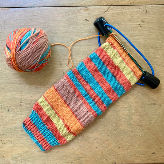 Handknit sock-in-progress, resting on a black plastic double point knitting needle holder with blue elastic. The sock and the working yarn are striped in shades of orange, yellow, and aqua blue.