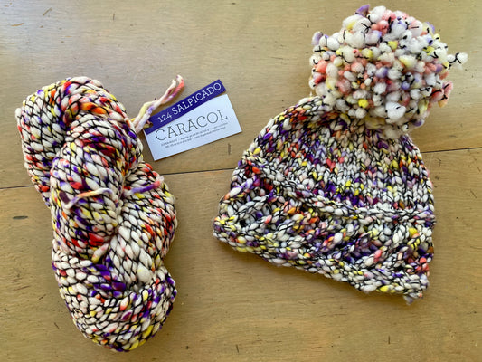 Speckled skein of Malabrigo Caracol yarn and a hat made in the same white, orange, and yellow speckled colorway lying on a wooden surface.