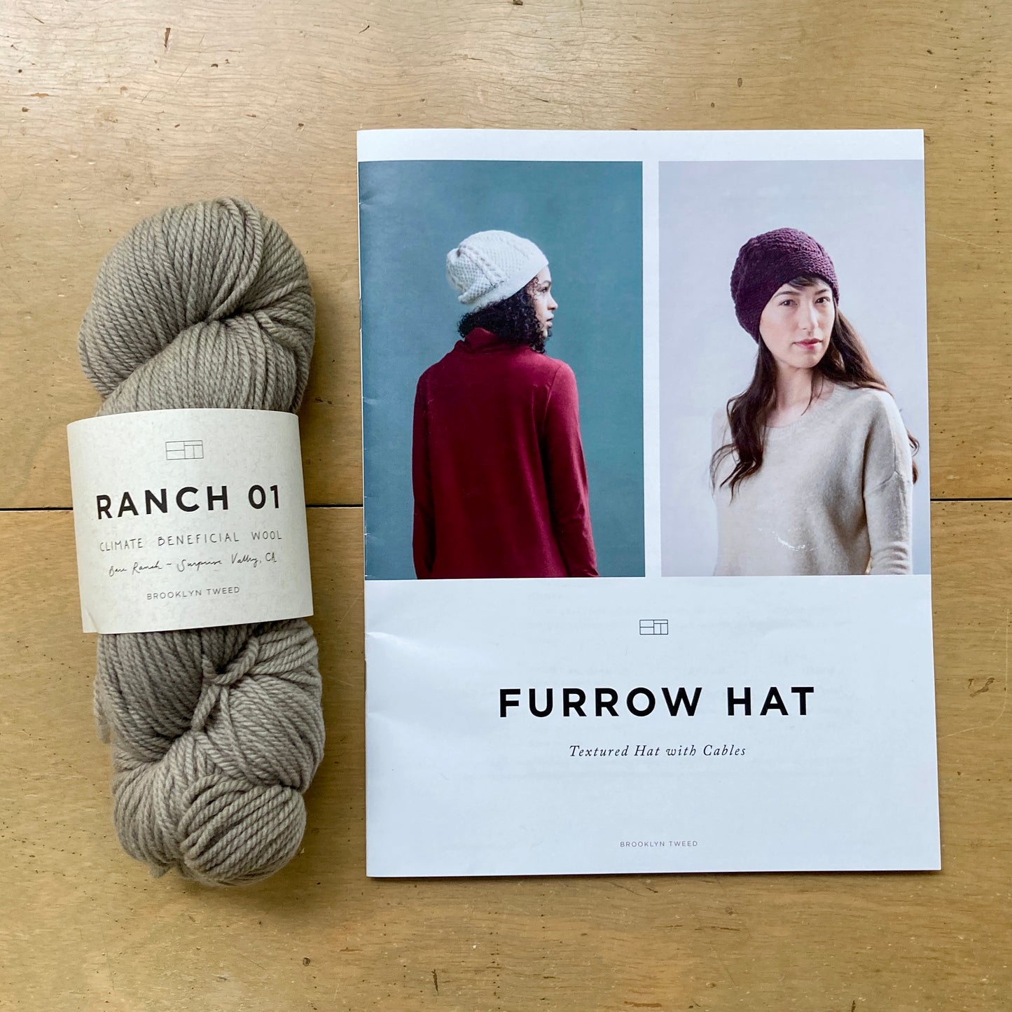 One skein of Brooklyn Tweed Ranch 01 yarn and a print copy of a hat pattern lie on a wooden surface.