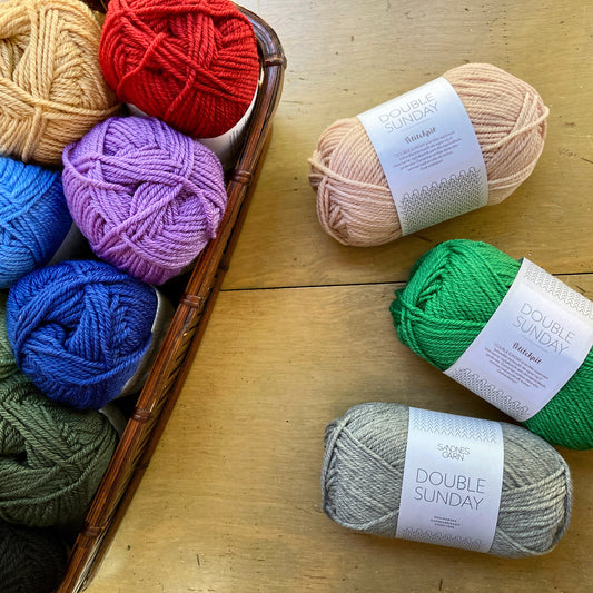 Colorful balls of Sandnes Garn Double Sunday in a rectangular basket and arranged on the wooden surface beside it.
