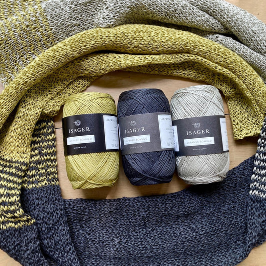 Handknit scarf arranged in a circle on a wooden surface, with 3 balls of Isager Japansk Bomuld yarn at the center.