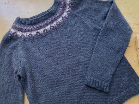 Dark blue grey sweater with a purple colorwork yoke lying on a wooden surface.