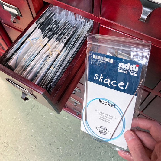 Close up on a hand holding Addi Rocket needles in front of a library card catalog with one drawer open, holding circular knitting needles.