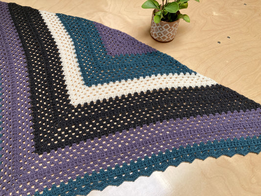 Triangular crocheted shawl lying flat on a table near a potted plant.