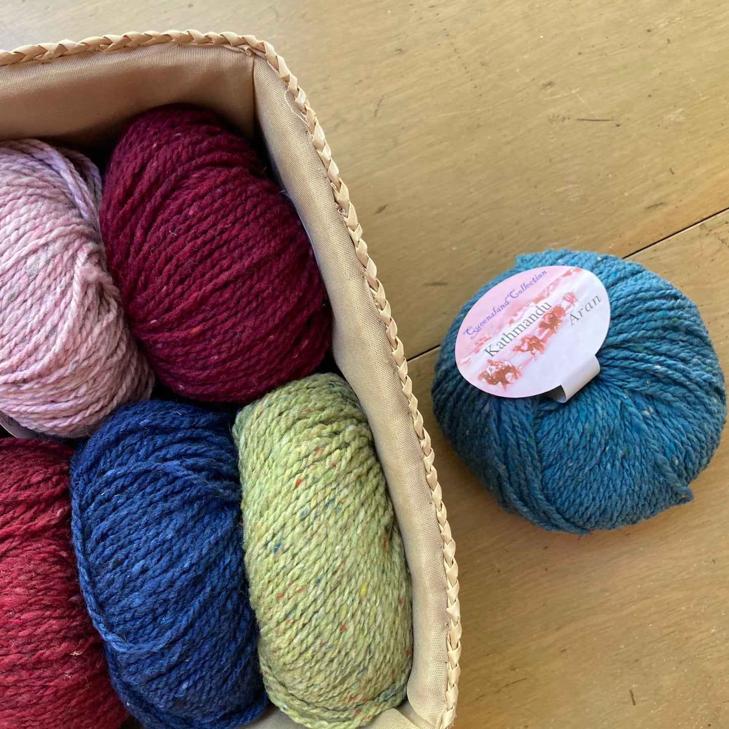 Colorful balls of tweed Queensland Kathmandu Aran yarn in a rectangular basket on a wooden surface.
