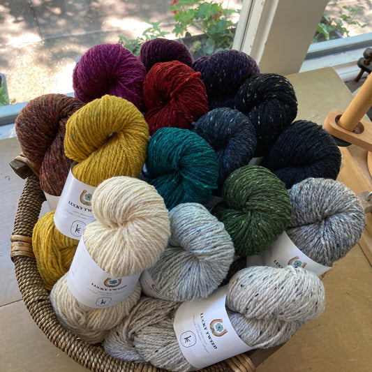 Colorful skeins of Kelbourne Woolens Lucky Tweed yarn in a round basket on a wooden table with a ballwinder.