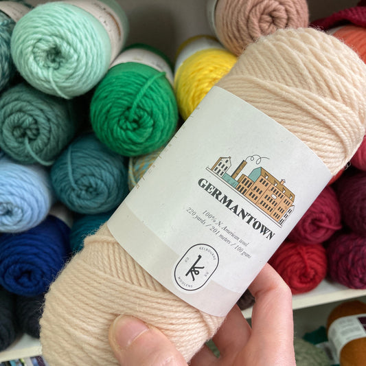 A hand holds up a skein of Kelbourne Woolens Germantown yarn before a shelf of colorful skeins.