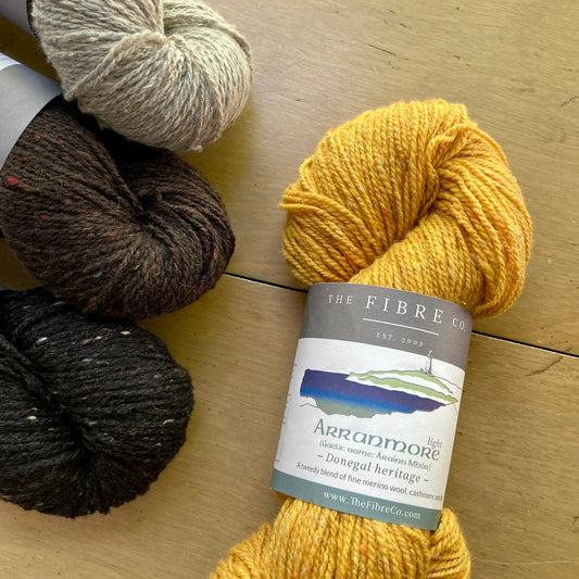 Four skeins of Fibre Co. Arranmore Light yarn in black, brown, beige, and yellow, laying on a wooden surface.