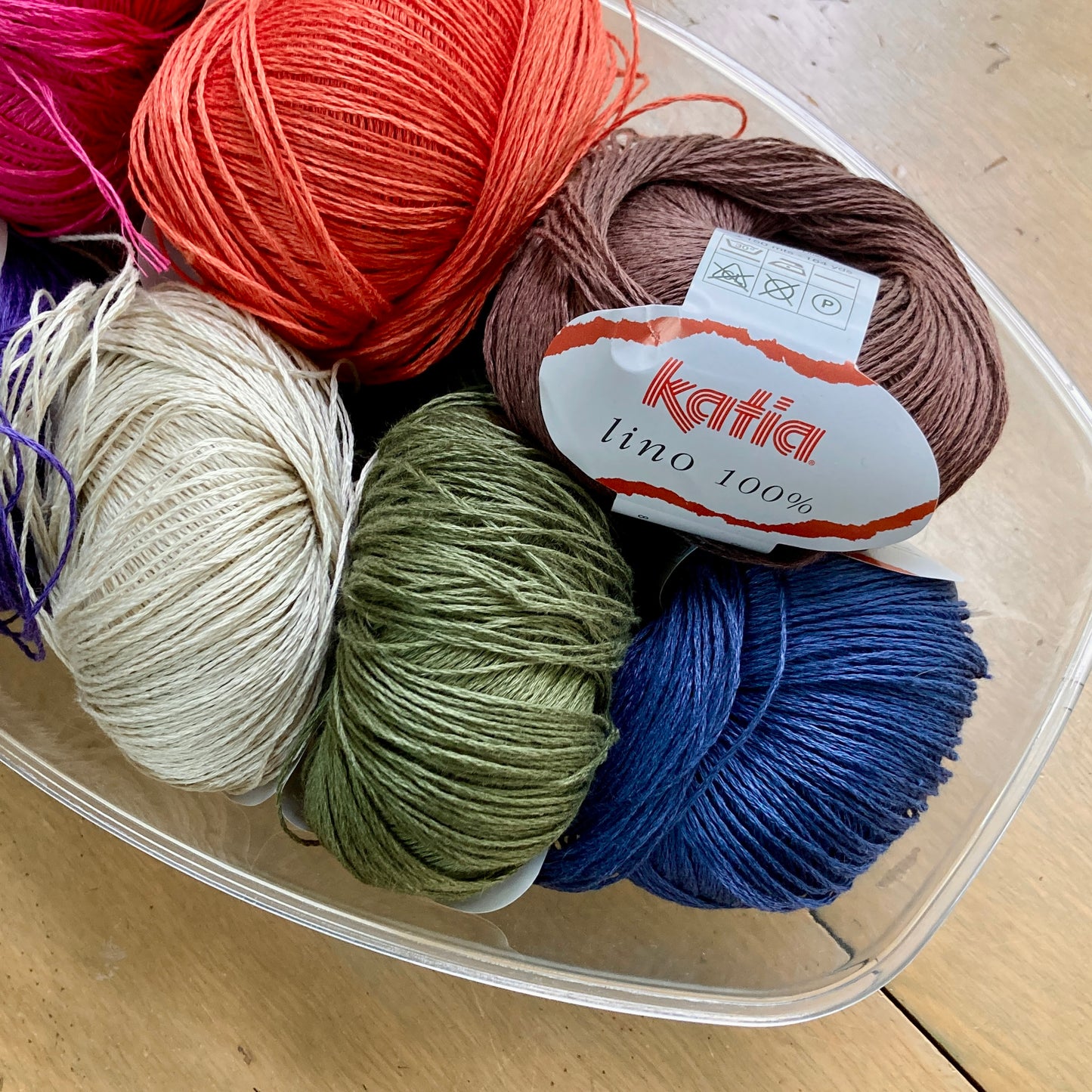 Colorful balls of Katia Lino yarn in a clear bowl on a wooden surface.