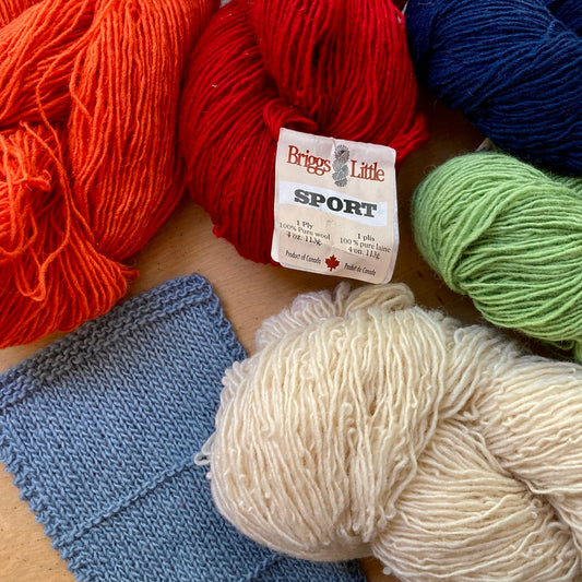 Close up on colorful skeins of Briggs & Little Sport yarn, arranged around a blue gray handknit swatch.