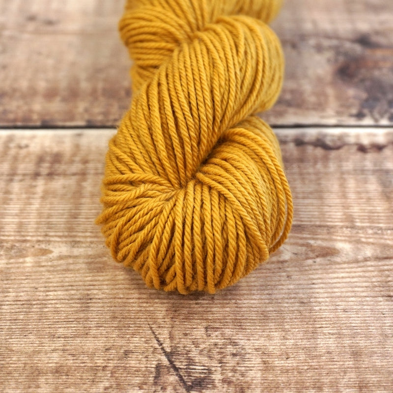 Mustard yellow skein of CoopKnits Socks Yeah! DK yarn on a wooden surface.