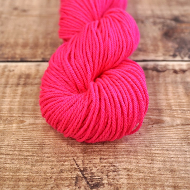Neon pink skein of CoopKnits Socks Yeah! DK yarn on a wooden surface.