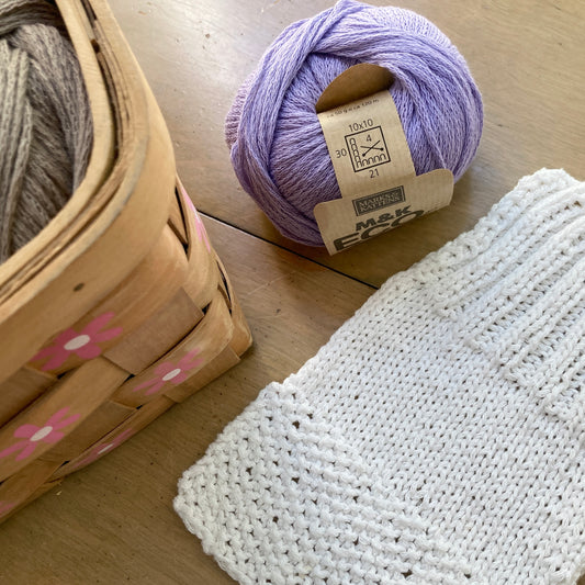 Lavender ball of M&K Eco Bomull yarn lying beside a white handknit swatch and a floral print basket, all arranged on a wooden surface.