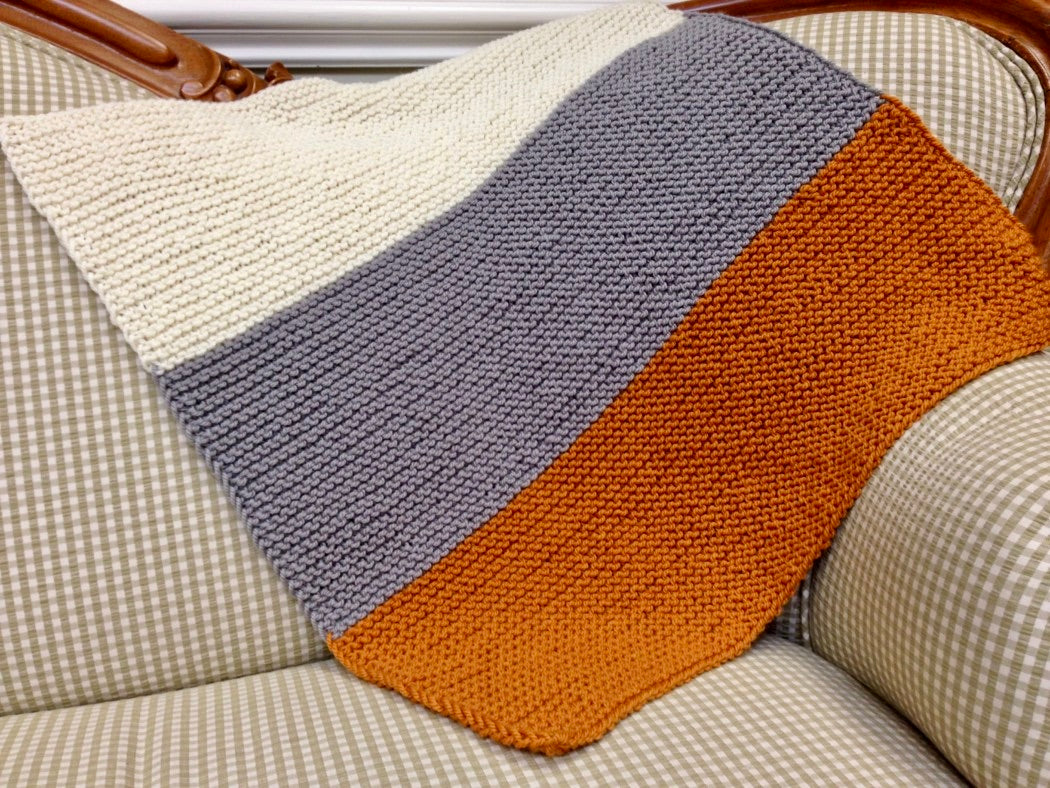Colorblock garter stitch blanket in cream, gray, and orange, draped on a gingham sofa on display at the Hillsborough Yarn Shop.
