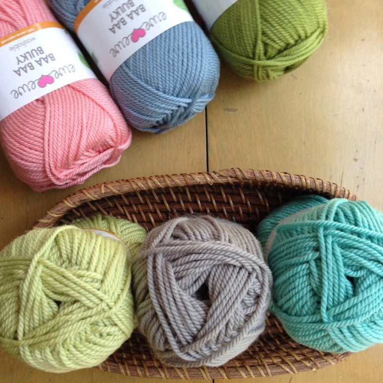 Balls of pastel Ewe Ewe Baa Baa Bulky yarn in a basket and lying on a wooden surface.