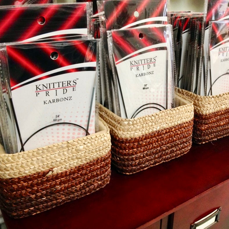 Small rectangular baskets holding Knitter’s Pride Karbonz circular needles, sitting on top of a dark burgundy library card catalog.