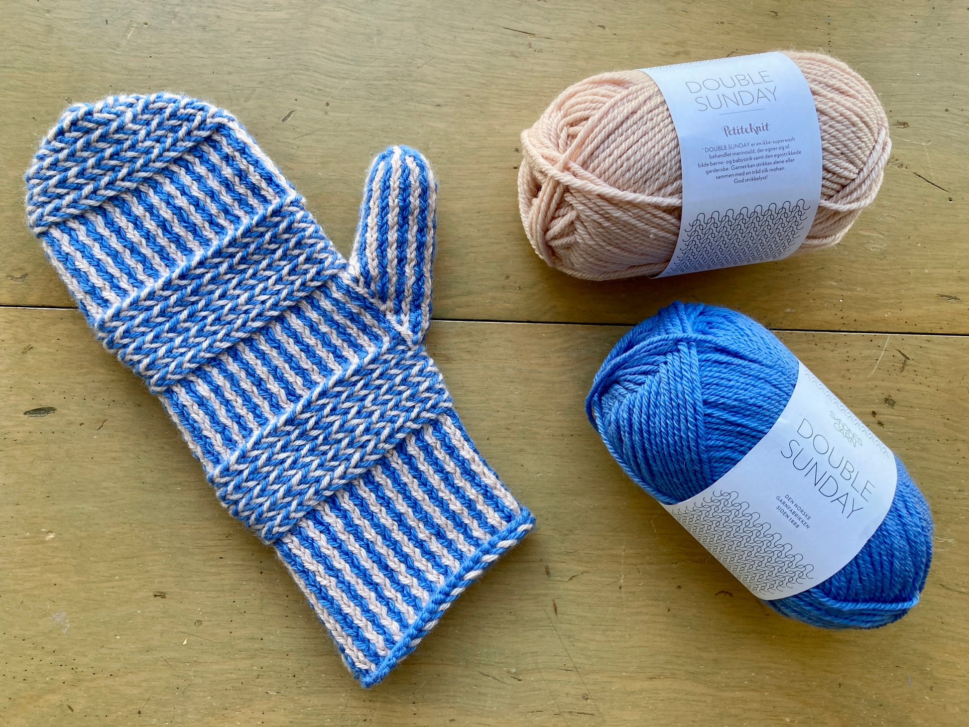 Blue and pink colorwork mitten lying on a wooden surface next to blue and pink balls of Sandnes Garn Double Sunday yarn.