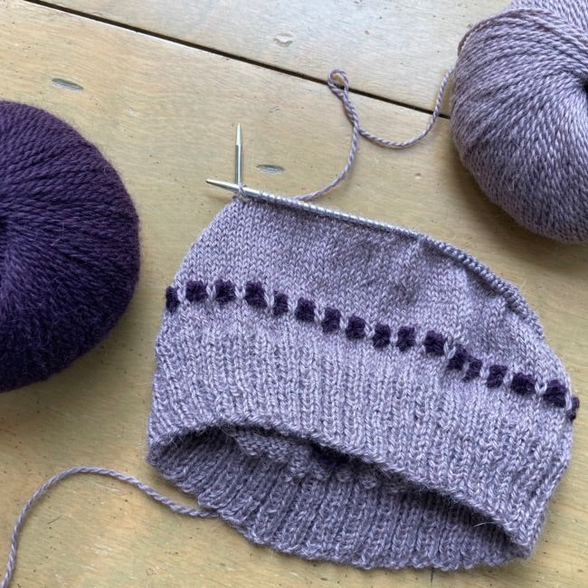 Lavender and dark purple hat in progress, lying between two balls of yarn on a wooden surface.