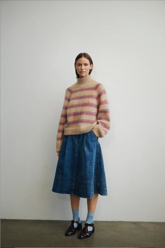 Model stands against white backdrop wearing the Norma sweater, designed by My Favourite Things Knitwear as part of Isager Archives collection, in sand color with light blue and red stripe motif.