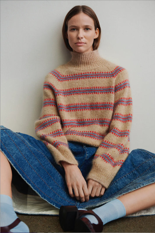Model sitting against white backdrop wearing the Norma sweater, designed by My Favourite Things Knitwear as part of Isager Archives collection, in sand color with light blue and red stripe motif.