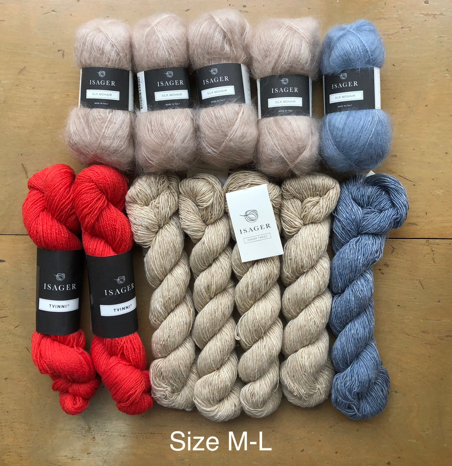 Contents of M-L Norma sweater kit on wooden table. Clockwise from left: 4 balls Isager Silk Mohair in beige, one ball Isager Silk Mohair light blue, one skein Isager Tweed in Topaz, 4 skeins Isager Tweed in Sand, and two skeins Isager Tvinni in red.