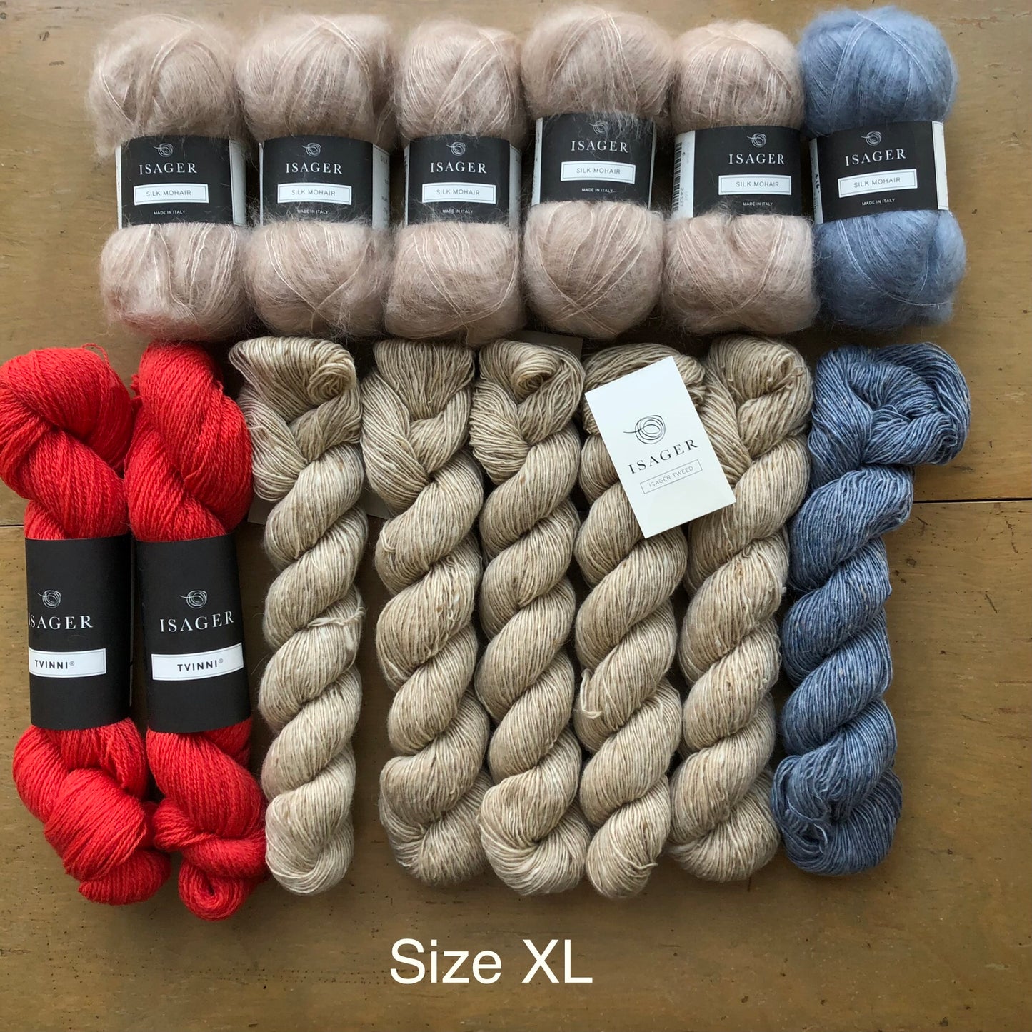 Contents of XL Norma sweater kit on wooden table. Clockwise from left: 5 balls Isager Silk Mohair in beige, one ball Isager Silk Mohair light blue, one skein Isager Tweed in Topaz, 5 skeins Isager Tweed in Sand, and two skeins Isager Tvinni in red.
