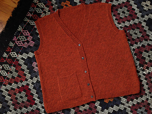 Deep rust handknit vest with buttons, lying flat on a rug with diamond motifs.