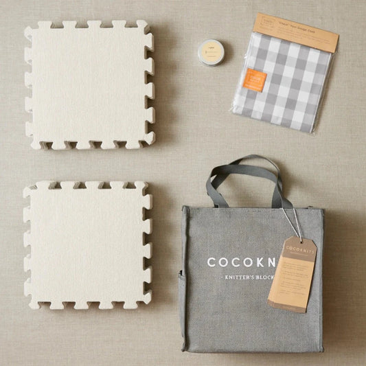 Square gray tiles with puzzle piece edges, a gray jute tote, a small round tin of t-pins, and a gray and white gingham cloth arranged on a beige surface.
