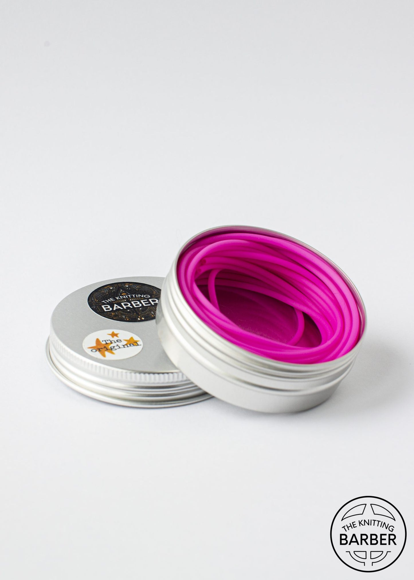 Round silver tin lying open beside its lid, showing pink Knitting Barber Cords coiled inside.