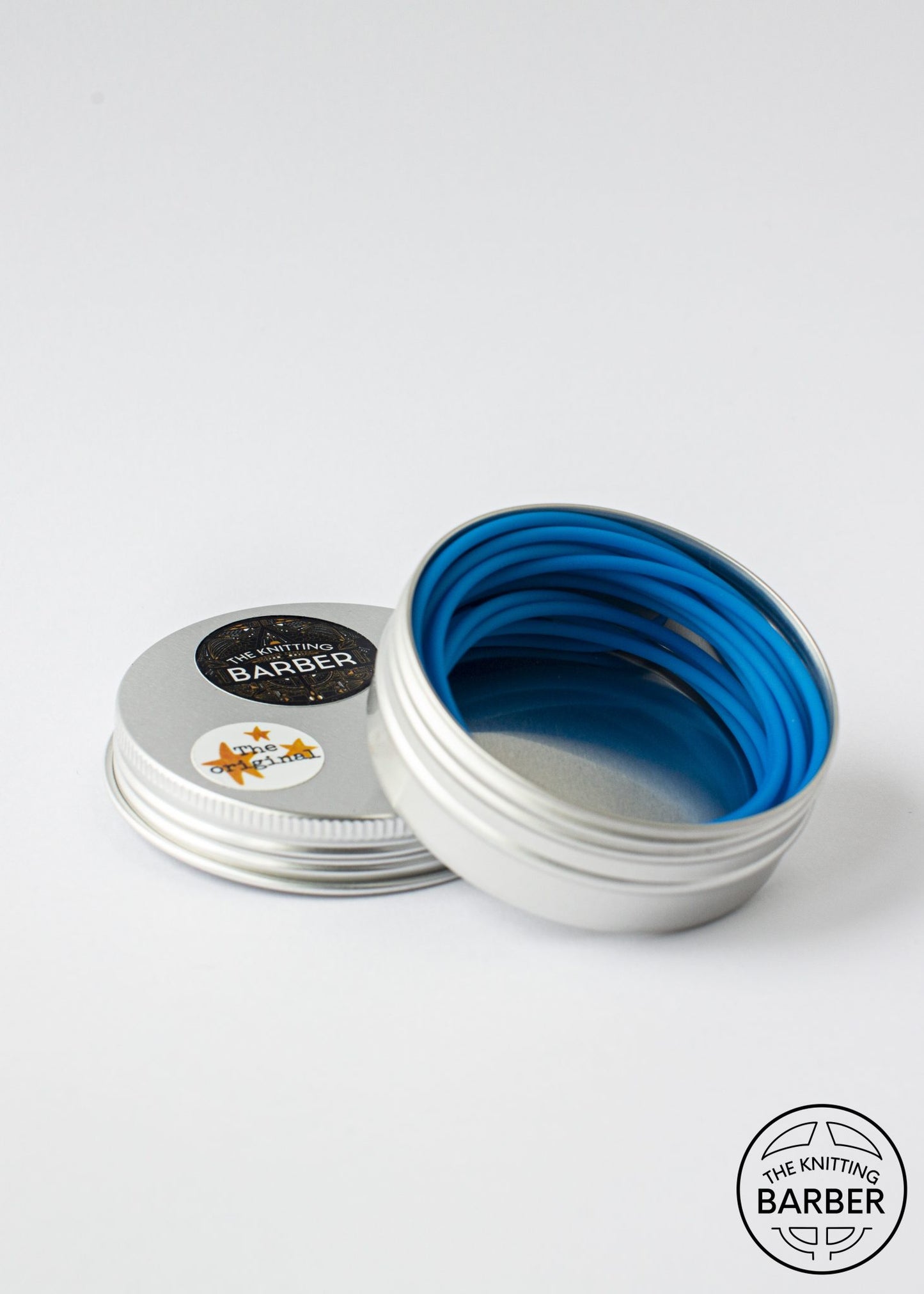 Round silver tin lying open beside its lid, showing light blue Knitting Barber Cords coiled inside.
