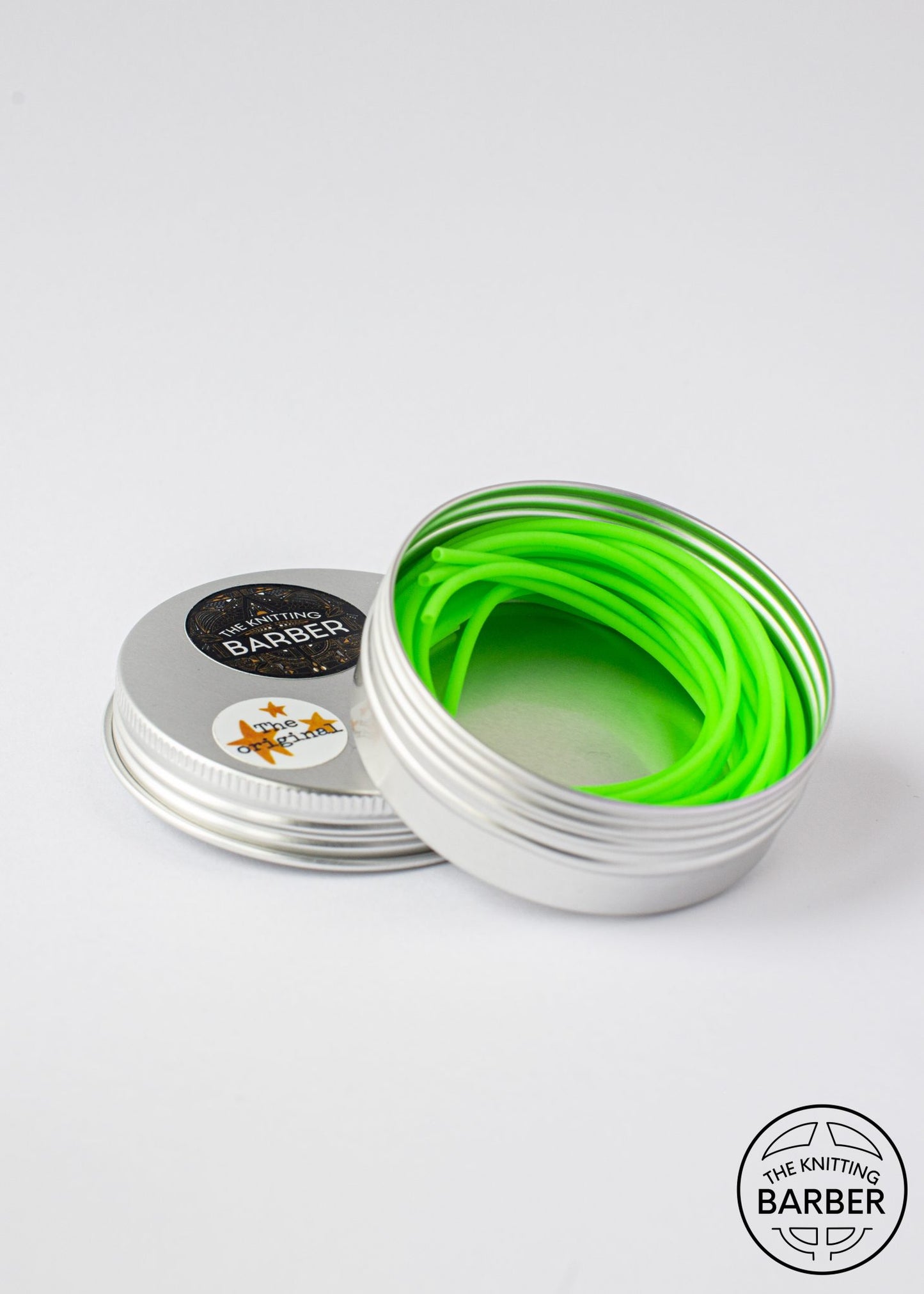 Round silver tin lying open beside its lid, showing bright green Knitting Barber Cords coiled inside.