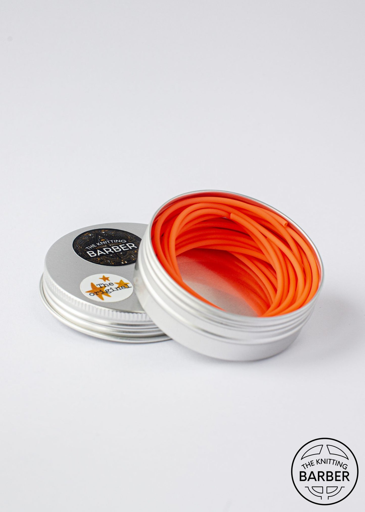 Round silver tin lying open beside its lid, showing orange Knitting Barber Cords coiled inside.