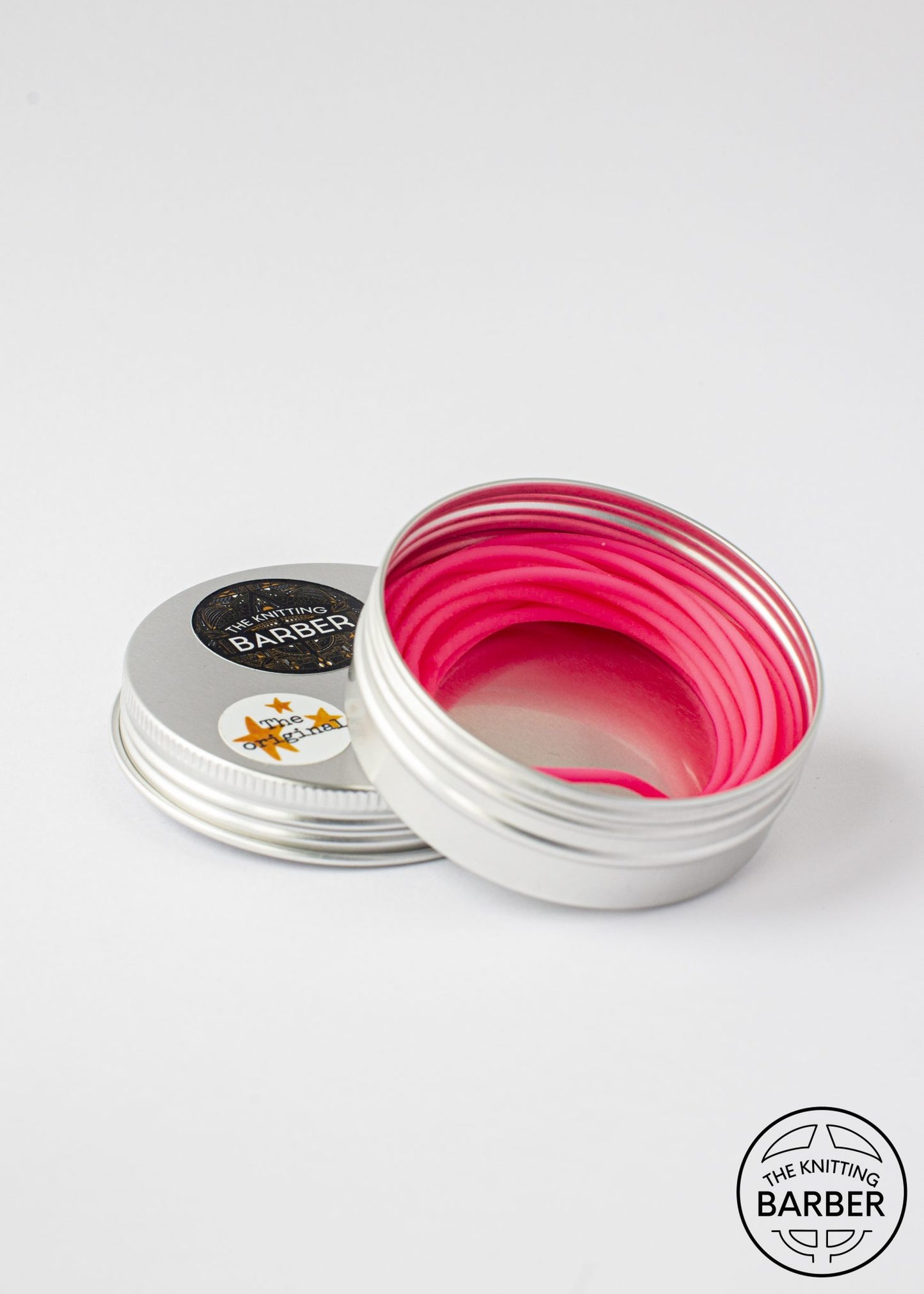 Round silver tin lying open beside its lid, showing pink Knitting Barber Cords coiled inside.