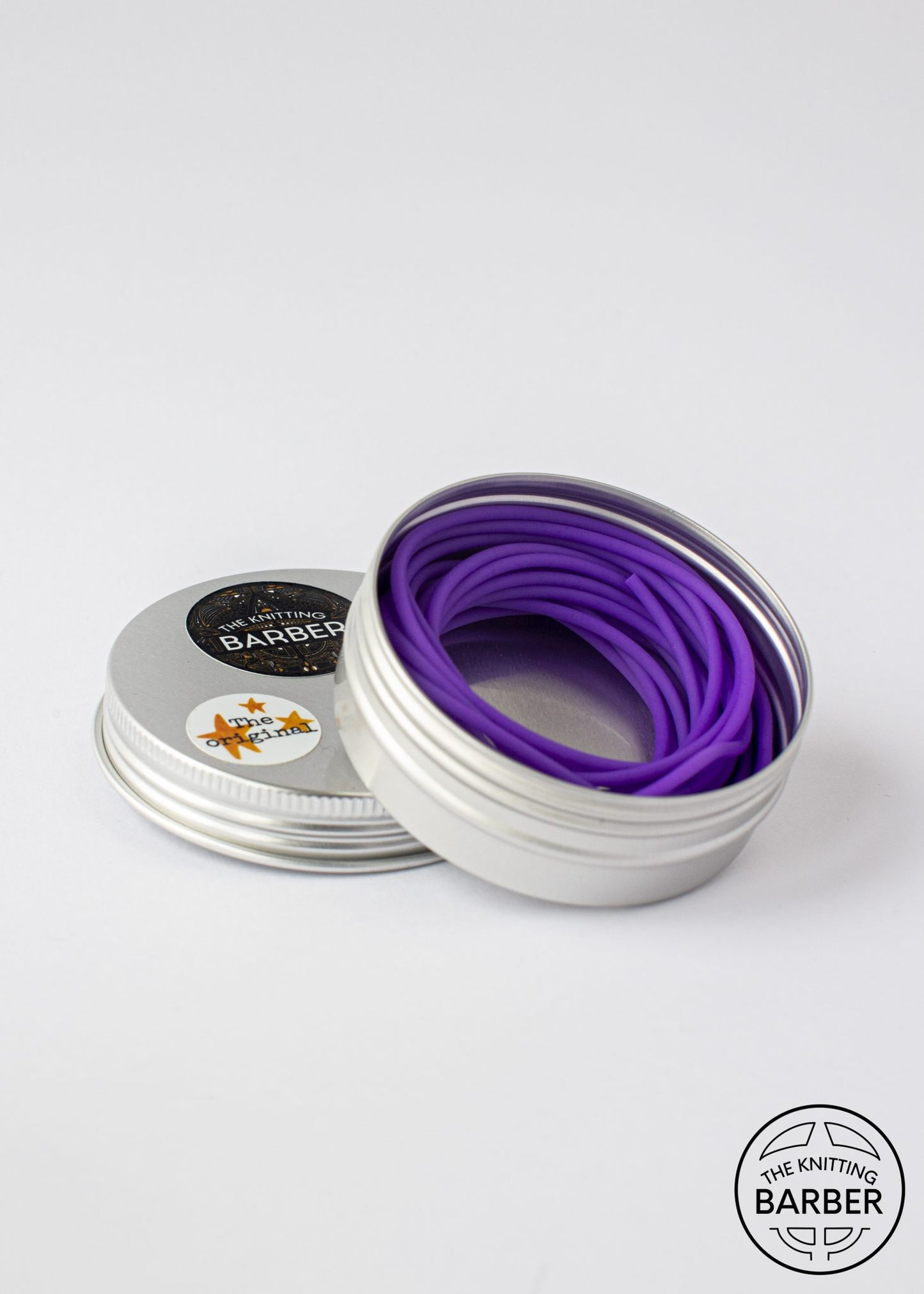 Round silver tin lying open beside its lid, showing purple Knitting Barber Cords coiled inside.