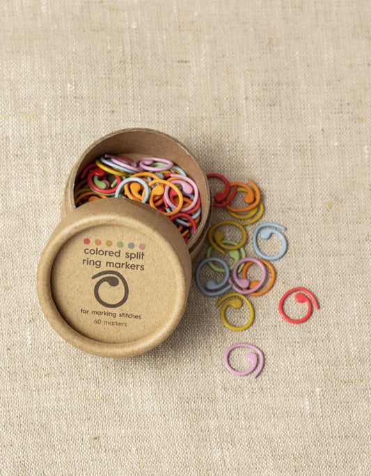 Open container showing colorful split ring stitch markers inside, and a few scattered beside the round kraft brown box.