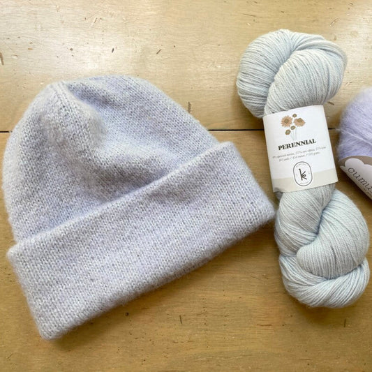 Pale gray PetiteKnit "Oslo Hat" on a wooden surface, next to skeins of Kelbourne Woolens Perennial and Fyberspates Cumulus yarn.