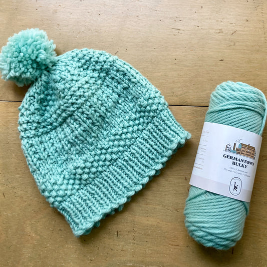 Mint green handknit hat next to a skein of yarn in the same color, lying on a wooden surface. 