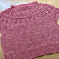 Pink handknit sweater with short sleeves and lace design at yoke, lying on a wooden surface on display at the Hillsborough Yarn Shop. 