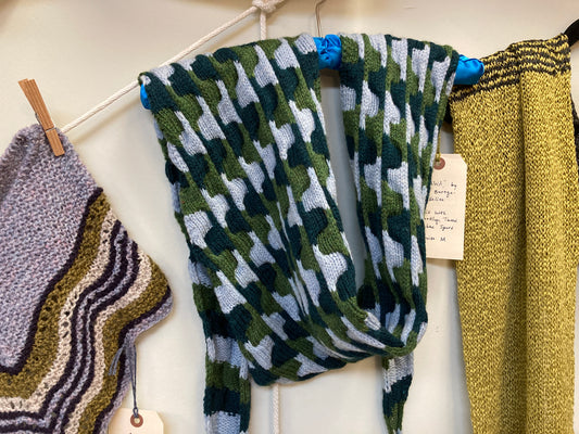 Textured handknit scarf in light blue, grass and forest green, hanging on a blue hanger on a white wall on display at the Hillsborough Yarn Shop. 