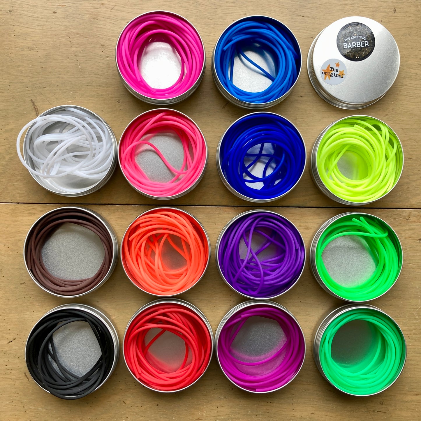 Small round tins with colorful Knitting Barber Cords coiled inside, arranged in a grid on a wooden surface. 