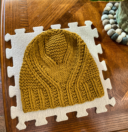 Handknit mustard yellow hat blocking on a light gray mat, lying on a wooden surface. 