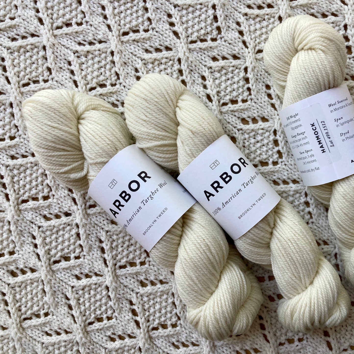 Three skeins of natural white Brooklyn Tweed Arbor yarn lying on a handknit lace blanket in the same color. 