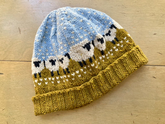 Handknit colorwork hat featuring a black and white sheep motif on a mustard yellow and sky blue background, lying on a wooden surface. 