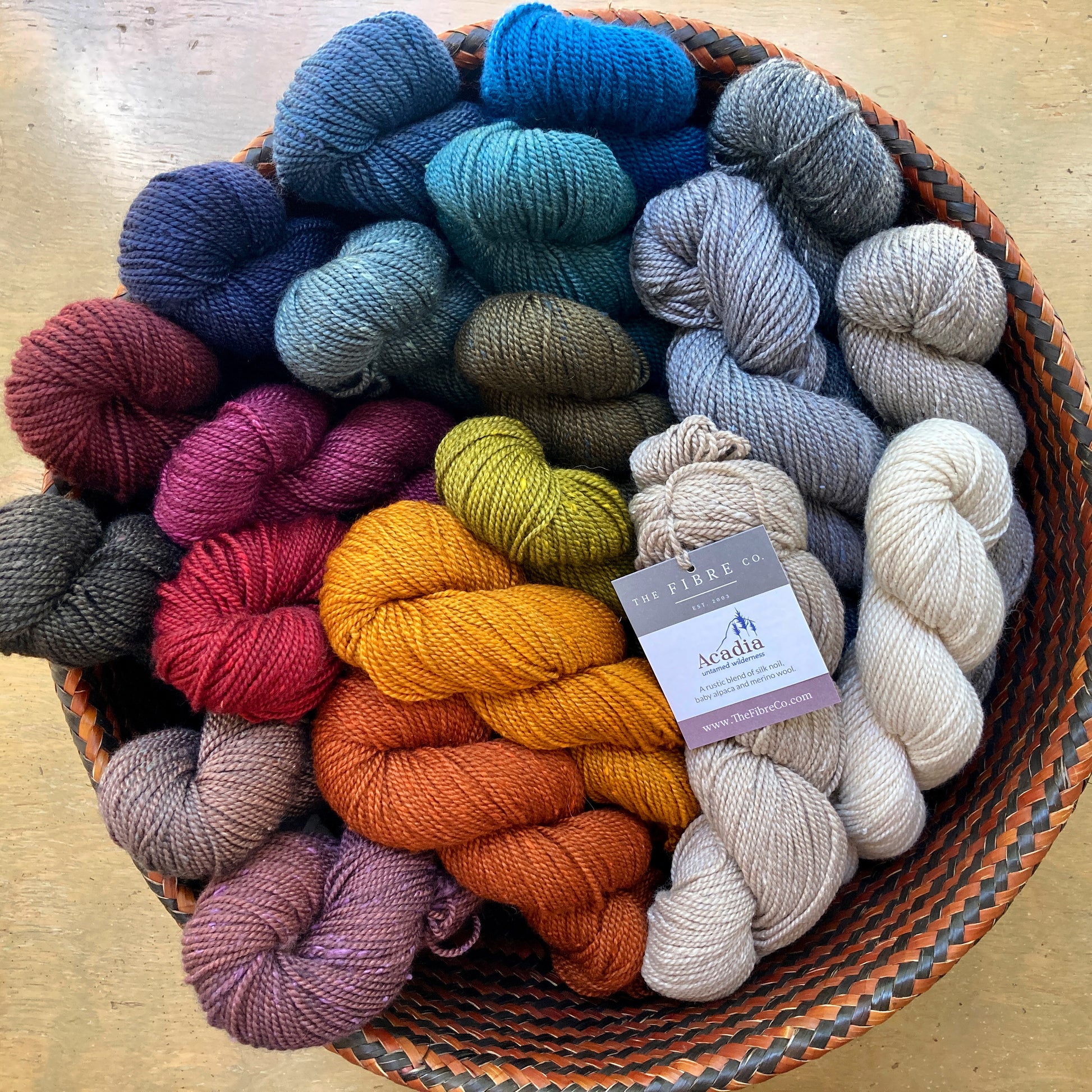 Basket of colorful skeins of Fibre Co. Acadia yarn sitting on a wooden table. 