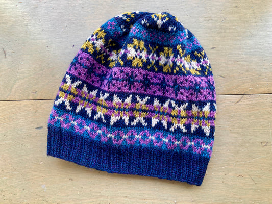 A fair isle hat in shades of blue, purple, and yellow lying on a wooden surface. 